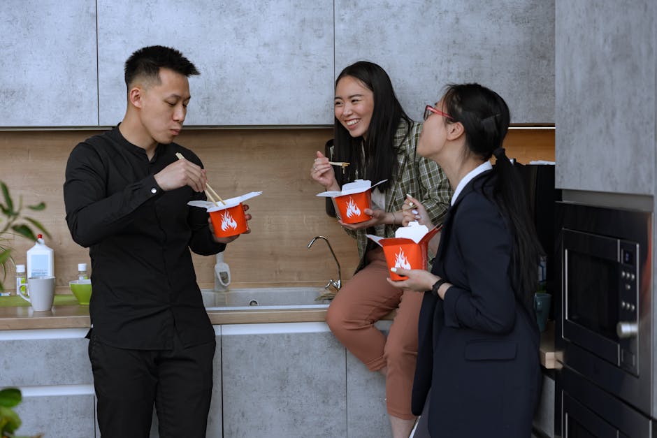 Three colleagues enjoy a casual lunch break with takeout food in an office kitchen.